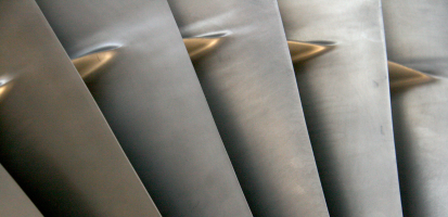 Close-up image of what appear to be titanium turbofan engine fan blades, showing their smooth curved surfaces, metallic gray finish, and soft reflections that highlight the aerodynamic shape and texture of the blades.