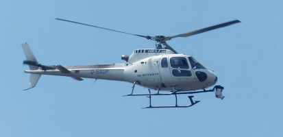White helicopter in flight against a clear blue sky with camera equipment mounted underneath.