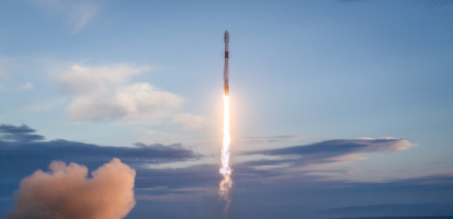 A white rocket launches vertically into a partly cloudy sky at sunrise or sunset, leaving a bright trail of flame and smoke behind it. The rocket’s silhouette is sharply outlined against the soft blue and orange hues of the sky, with a plume of exhaust expanding near the launch pad below.