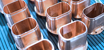 Close-up of polished, rectangular copper components with rounded edges arranged in rows on a blue perforated conveyor belt, highlighting their clean, reflective finish.