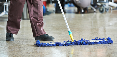 A person in brown trousers using a blue dust mop to clean a glossy floor in a commercial space, showing the mop in mid-sweep with the background slightly blurred to emphasize the cleaning action.